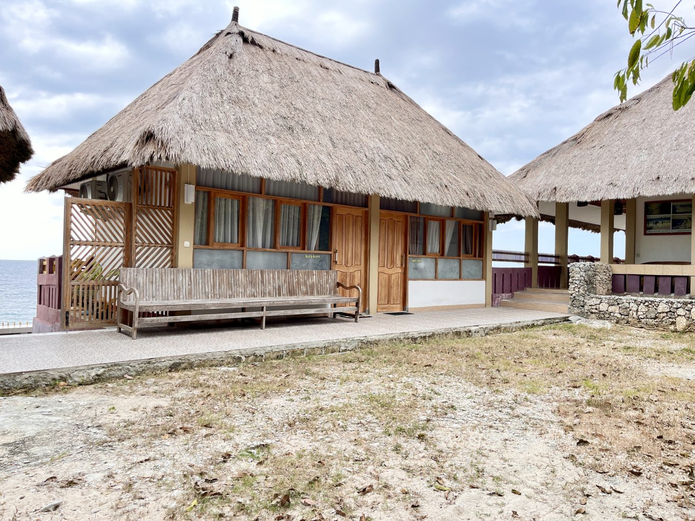 A traditional Redemptorist Villa building with a large thatched roof and wooden features sits by the ocean under a cloudy sky, with a long wooden bench on its porch.