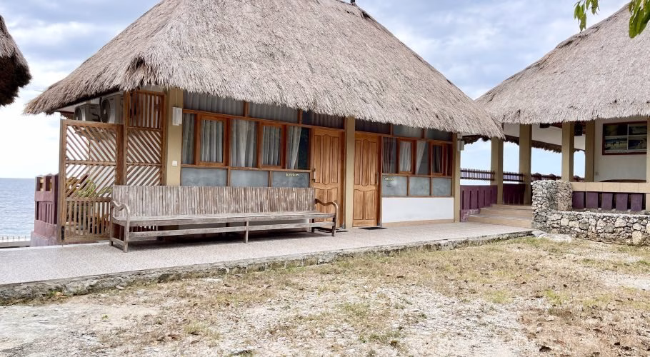 A traditional Redemptorist Villa building with a large thatched roof and wooden features sits by the ocean under a cloudy sky, with a long wooden bench on its porch.