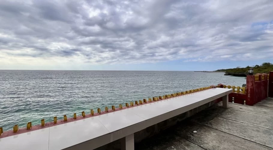 A wide ocean view under a cloudy sky, seen from a coastal patio with a long white bench and red and gold railing.