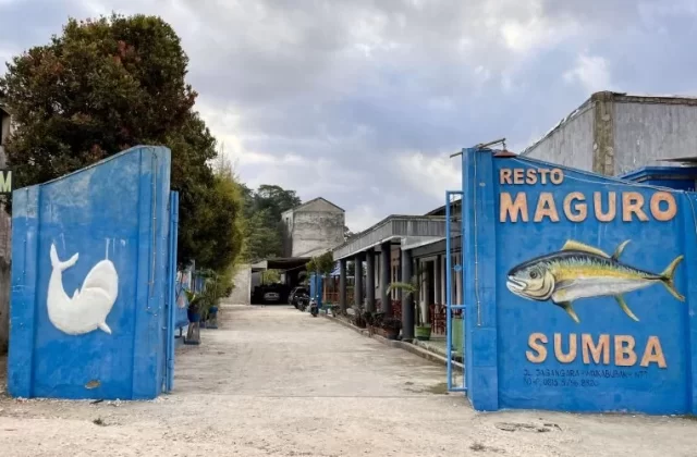 The blue entrance walls of Resto Maguro Sumba feature a large yellow fish and a white whale mural, leading to a restaurant complex under a cloudy sky.