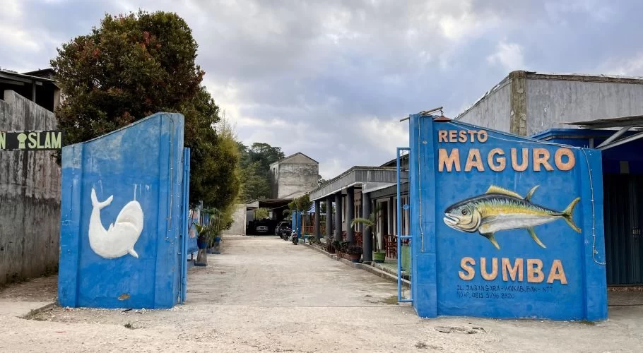 The blue entrance walls of Resto Maguro Sumba feature a large yellow fish and a white whale mural, leading to a restaurant complex under a cloudy sky.