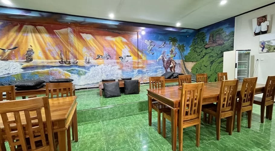 Restaurant interior with wooden tables and chairs on a green tiled floor, featuring a large mural depicting a vibrant seascape and forest.