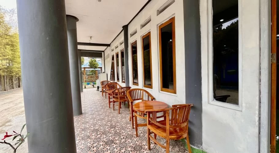 A long outdoor patio with dark grey pillars and white walls features several wooden tables and chairs on patterned tiles, next to a bamboo grove.
