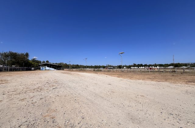 A wide, dry dirt horse racing track under a clear blue sky, with a building and trees left, and white spectator tents right.