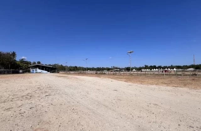 A wide, dry dirt horse racing track under a clear blue sky, with a building and trees left, and white spectator tents right.