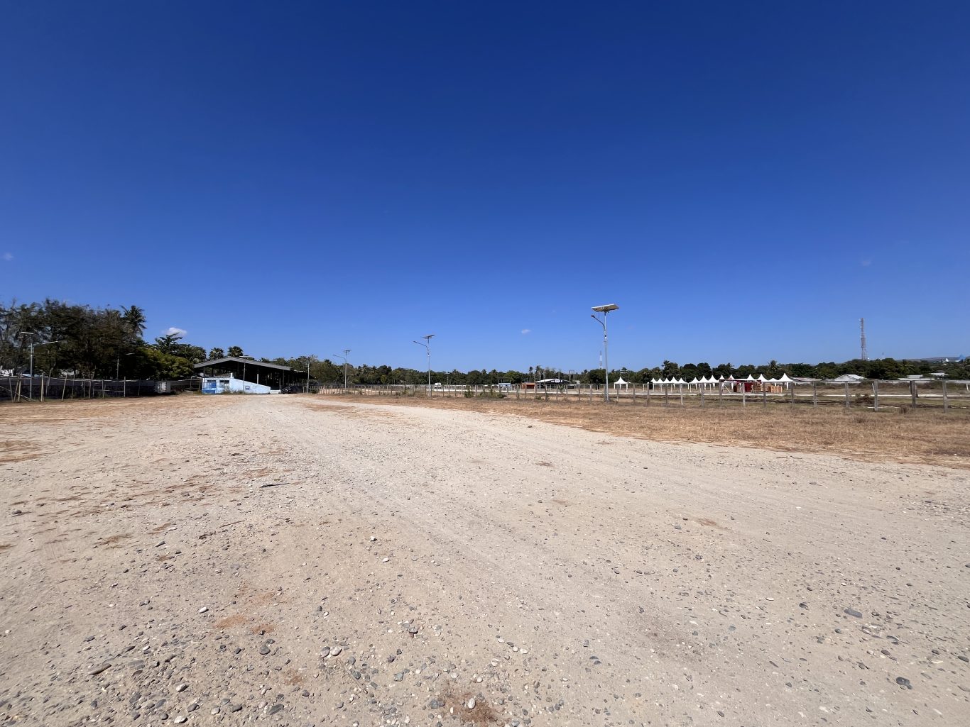 A wide, dry dirt horse racing track under a clear blue sky, with a building and trees left, and white spectator tents right.