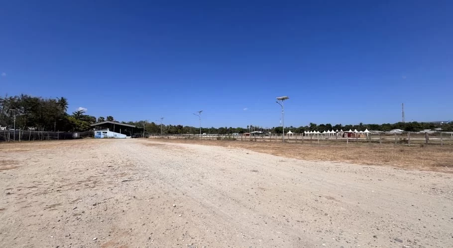 A wide, dry dirt horse racing track under a clear blue sky, with a building and trees left, and white spectator tents right.