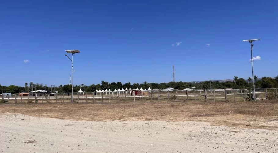 A dry, open field with a dirt track in the foreground, a fence, a row of white tents, and buildings under a clear blue sky, flanked by two solar streetlights.