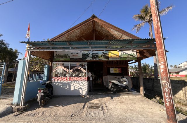 A modest, light-brown building with a peaked roof and a small awning, housing RM Mutiara Minang, under a clear blue sky. Two motorbikes are parked in front, and an Indonesian flag flies on the left.