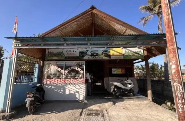 A modest, light-brown building with a peaked roof and a small awning, housing RM Mutiara Minang, under a clear blue sky. Two motorbikes are parked in front, and an Indonesian flag flies on the left.