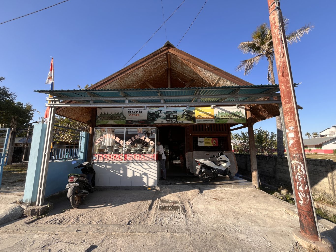 A modest, light-brown building with a peaked roof and a small awning, housing RM Mutiara Minang, under a clear blue sky. Two motorbikes are parked in front, and an Indonesian flag flies on the left.