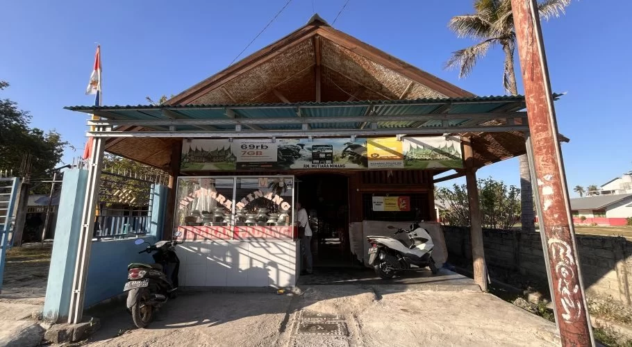 A modest, light-brown building with a peaked roof and a small awning, housing RM Mutiara Minang, under a clear blue sky. Two motorbikes are parked in front, and an Indonesian flag flies on the left.
