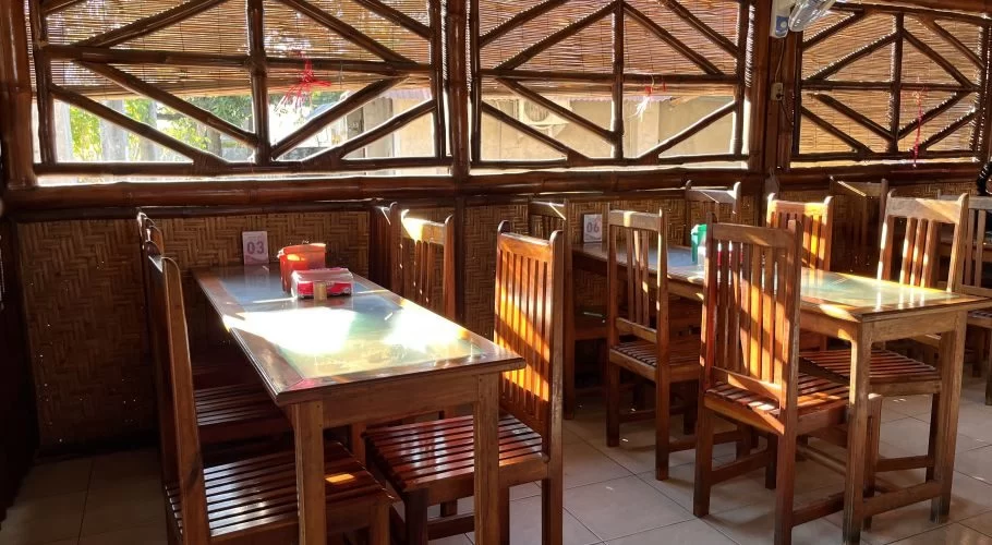 A tranquil restaurant interior features wooden tables and chairs, woven bamboo walls, and sunlit windows, creating a warm, earthy ambiance.