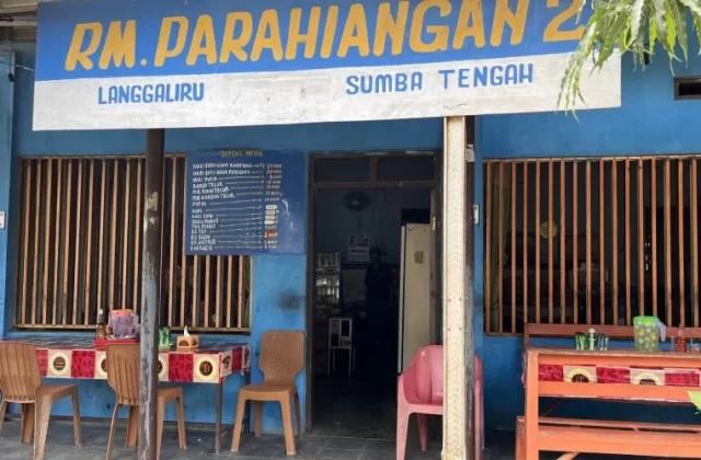 The blue exterior of RM Parahiangan 2 restaurant in Sumba Tengah features a yellow and white sign, outdoor tables with patterned cloths, and brown chairs.