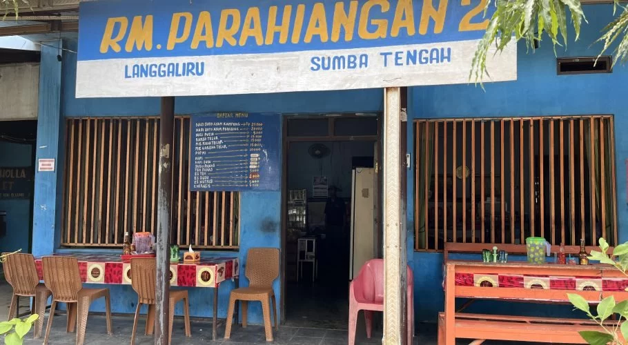 The blue exterior of RM Parahiangan 2 restaurant in Sumba Tengah features a yellow and white sign, outdoor tables with patterned cloths, and brown chairs.
