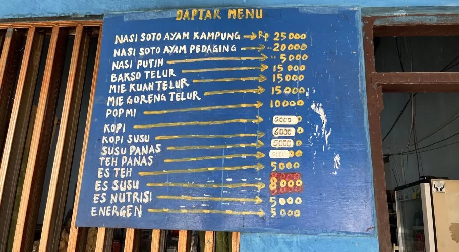 A worn blue wooden menu board displays handwritten food and drink items with prices in yellow and white text, partially covering wooden slats.