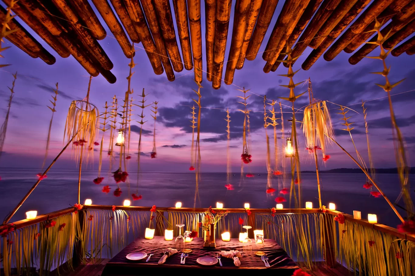 A romantic candlelit dining table for two is set under a bamboo canopy, overlooking a purple and pink ocean at dusk.