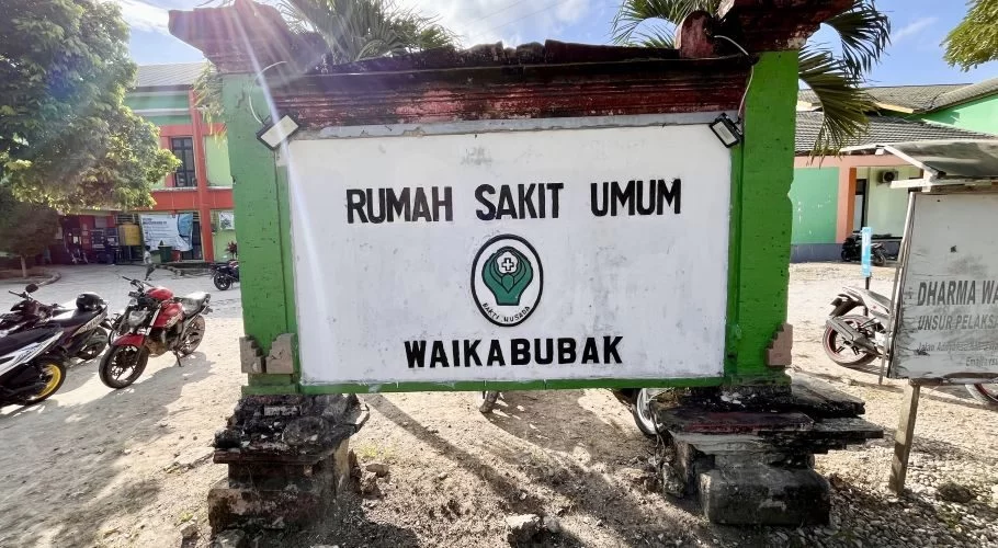 A green and white sign for "Rumah Sakit Umum Waikabubak" stands in front of a hospital building with parked motorcycles on a sunny day.