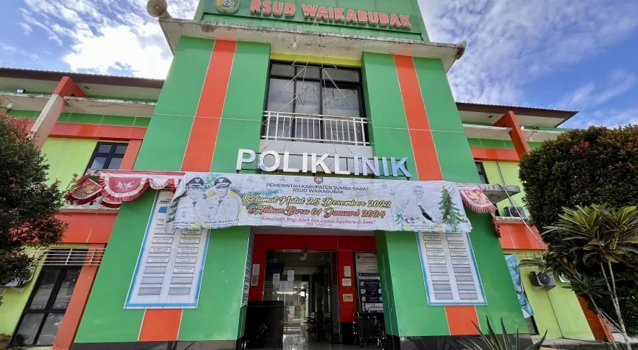 The RSUD Waikabubak hospital building in West Sumba features a green and orange striped facade with a 'POLIKLINIK' sign and a holiday banner.
