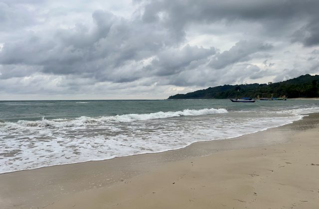 Gentle waves break on a sandy beach, with a small fishing boat in the calm ocean, backed by a lush green hill under a cloudy sky.