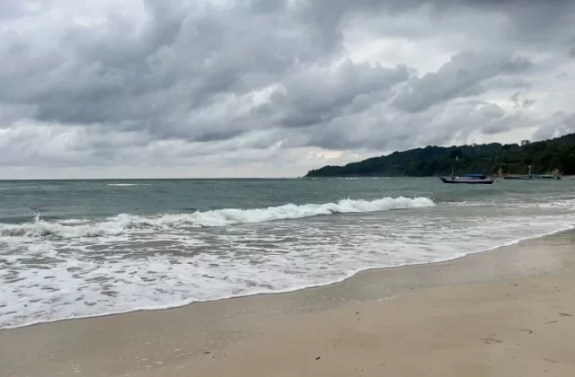 Gentle waves break on a sandy beach, with a small fishing boat in the calm ocean, backed by a lush green hill under a cloudy sky.
