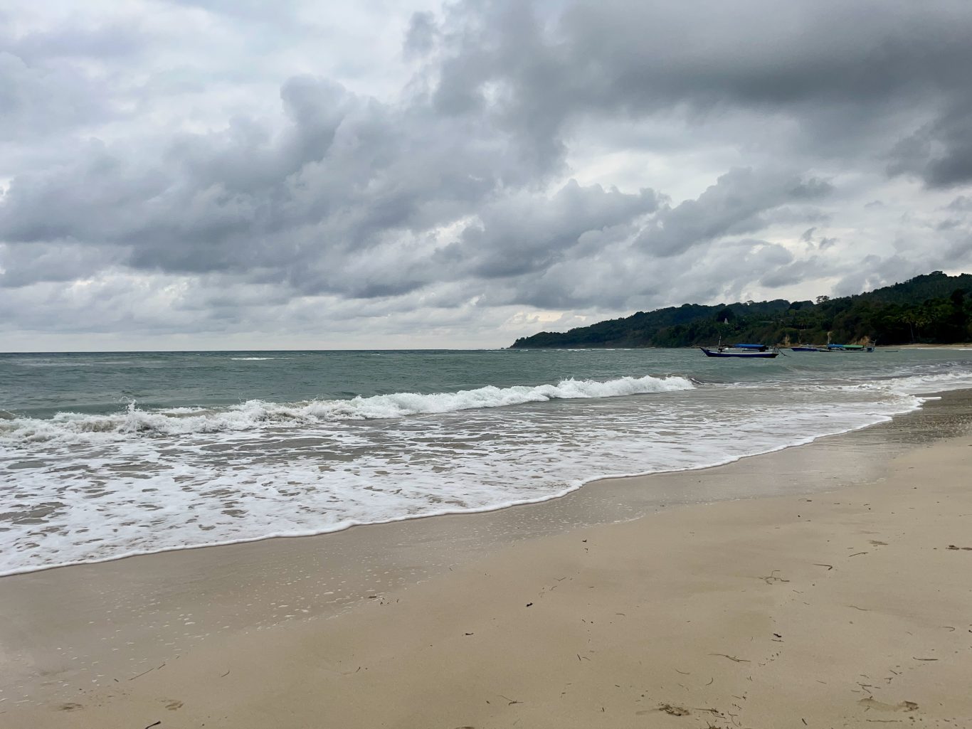 Gentle waves break on a sandy beach, with a small fishing boat in the calm ocean, backed by a lush green hill under a cloudy sky.