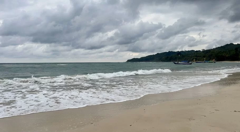 Gentle waves break on a sandy beach, with a small fishing boat in the calm ocean, backed by a lush green hill under a cloudy sky.