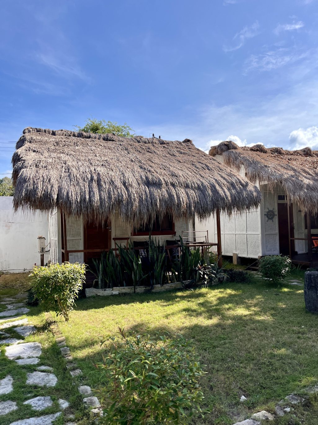 Two traditional Sumba resort bungalows with thick thatched roofs and light-colored woven walls stand under a blue sky, surrounded by green plants.