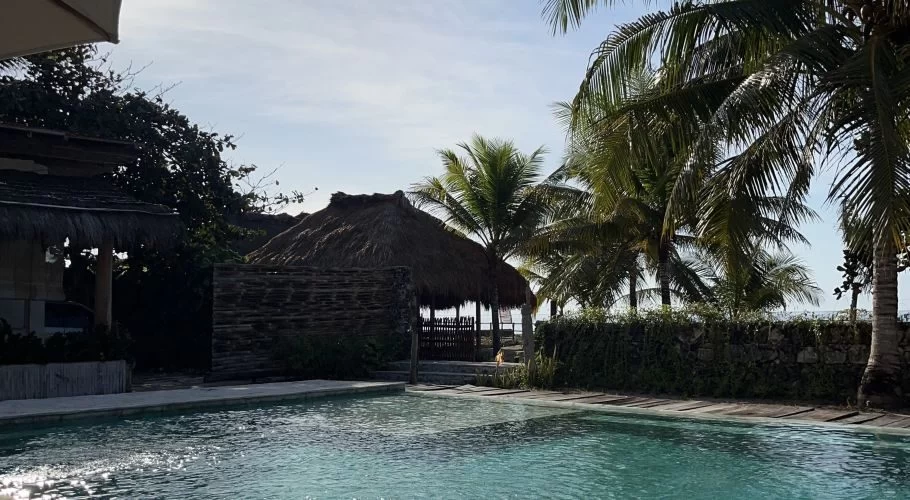 A turquoise swimming pool reflects sunlight, surrounded by lush palm trees and traditional thatched-roof buildings under a bright sky at Rua Beach Resort.
