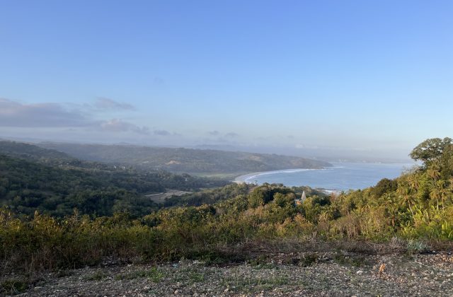Panoramic view from Rua Hill shows a curved sandy beach and blue ocean, bordered by lush green hills and valleys under a light blue sky.