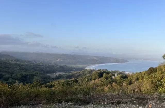 Panoramic view from Rua Hill shows a curved sandy beach and blue ocean, bordered by lush green hills and valleys under a light blue sky.