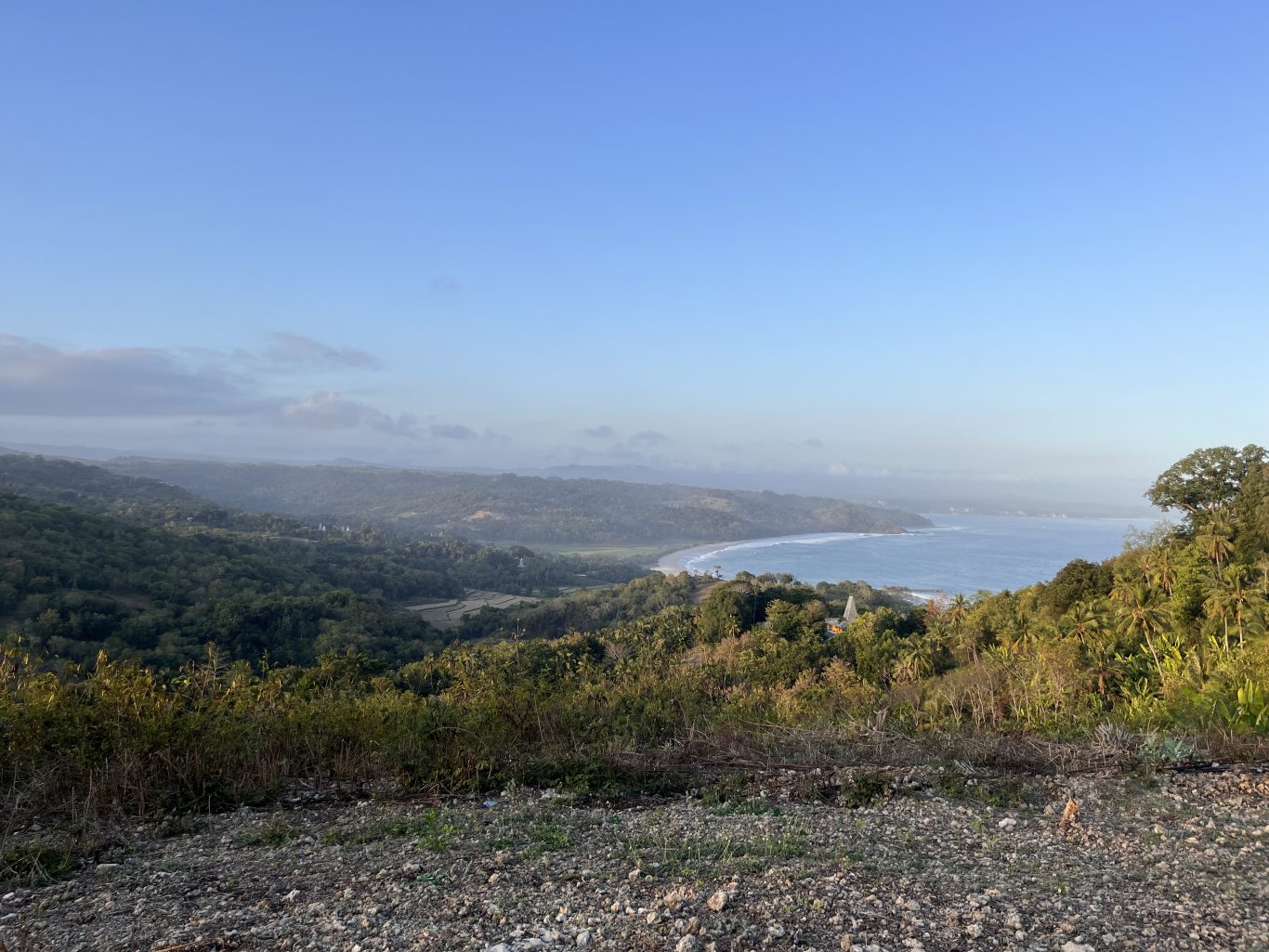 Panoramic view from Rua Hill shows a curved sandy beach and blue ocean, bordered by lush green hills and valleys under a light blue sky.