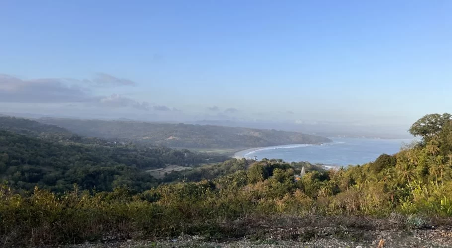 Panoramic view from Rua Hill shows a curved sandy beach and blue ocean, bordered by lush green hills and valleys under a light blue sky.