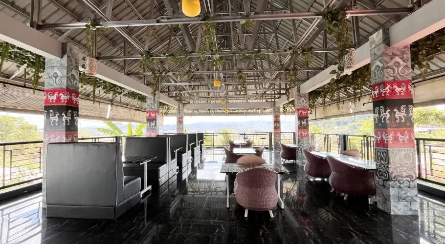 Open-air restaurant dining area with black booths, wicker chairs, traditional patterned pillars, and hanging greenery, overlooking green fields.