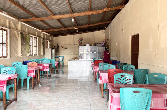 Interior of a simple restaurant with rows of teal and red plastic chairs at tables with pink tablecloths, a service counter, and a person in a maroon shirt.