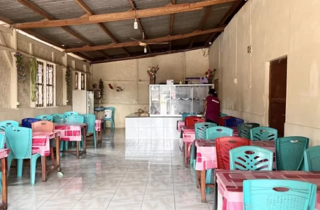 Interior of a simple restaurant with rows of teal and red plastic chairs at tables with pink tablecloths, a service counter, and a person in a maroon shirt.