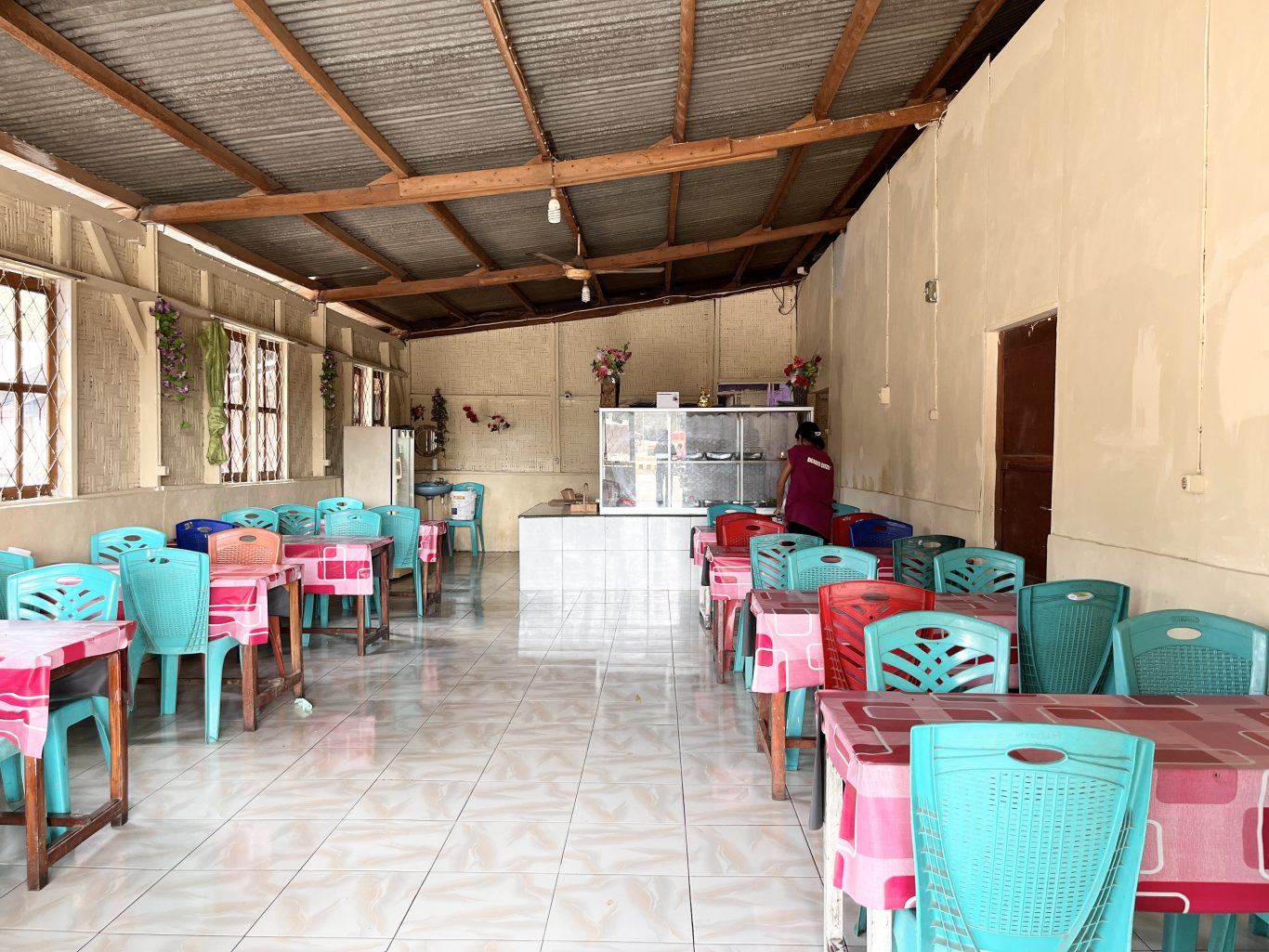 Interior of a simple restaurant with rows of teal and red plastic chairs at tables with pink tablecloths, a service counter, and a person in a maroon shirt.