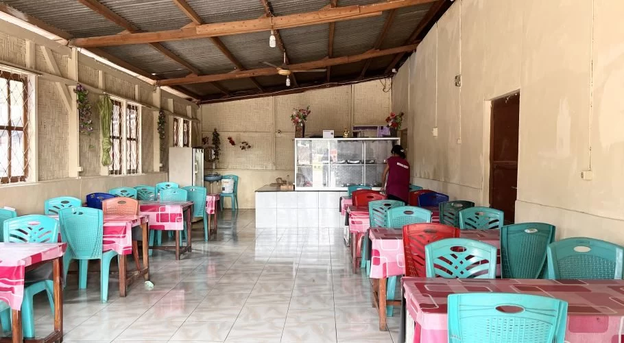 Interior of a simple restaurant with rows of teal and red plastic chairs at tables with pink tablecloths, a service counter, and a person in a maroon shirt.