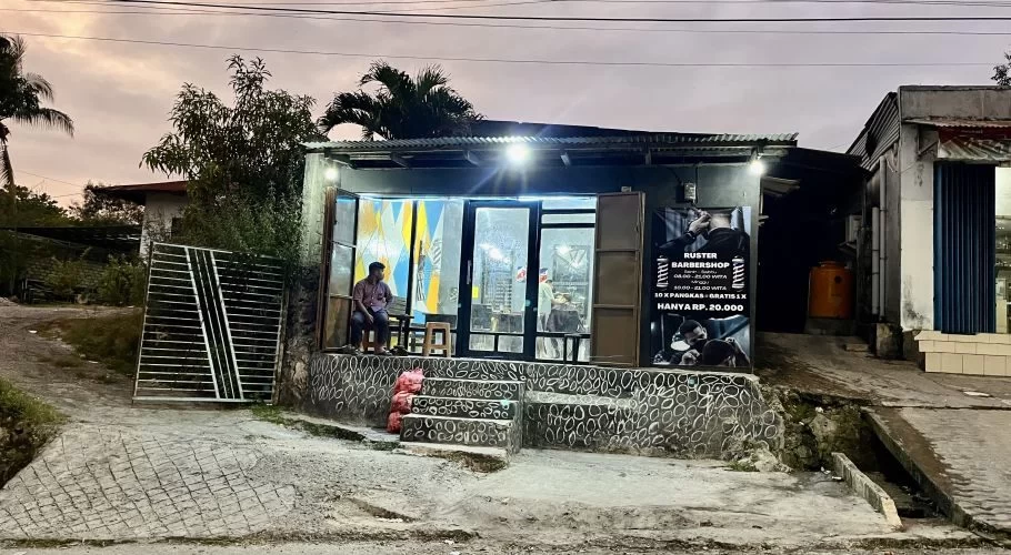 A man waits outside the brightly lit Ruster Barber Shop in Tambolaka, Sumba, under a cloudy evening sky. The shop has a corrugated roof and a large poster on its right side.