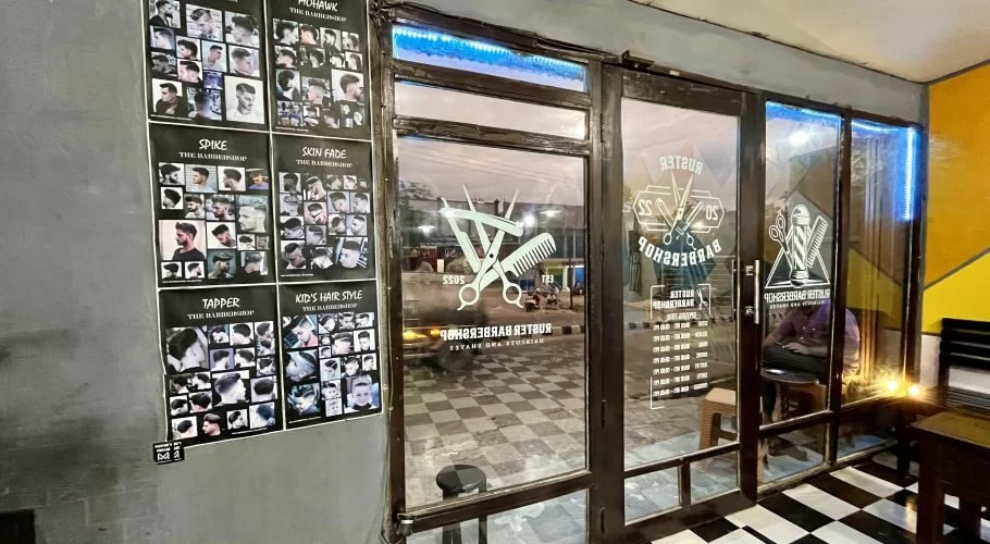 The modern exterior of Ruster Barber Shop in Tambolaka, Sumba, features glass doors with barber tool decals, blue LED lights, and posters displaying various hairstyles on a grey wall.