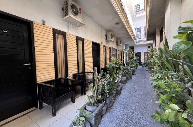 A narrow outdoor corridor at Sacca Residence homestay features black doors, light brown walls, green potted plants, and wooden benches.