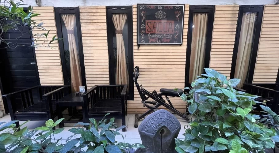 The exterior of Sacca Residence features a light wooden slatted wall, dark-framed doors, and a patio with dark wooden chairs and lush green plants.