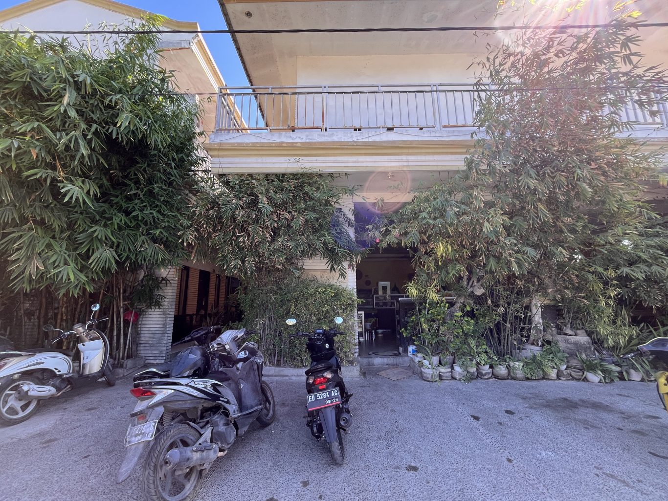 Sacca Restaurant's light-colored exterior with a balcony is framed by lush green bamboo and potted plants. Motorbikes are parked on the paved area.