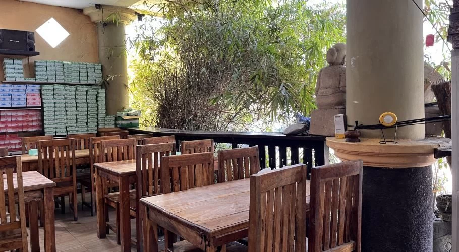 A spacious restaurant interior features numerous wooden tables and chairs, with stacks of boxes on shelves and green bamboo visible outside.