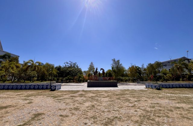 A monument featuring three dark horse statues stands prominently in Sandalwood Park, surrounded by green trees and dry grass under a bright blue sky.