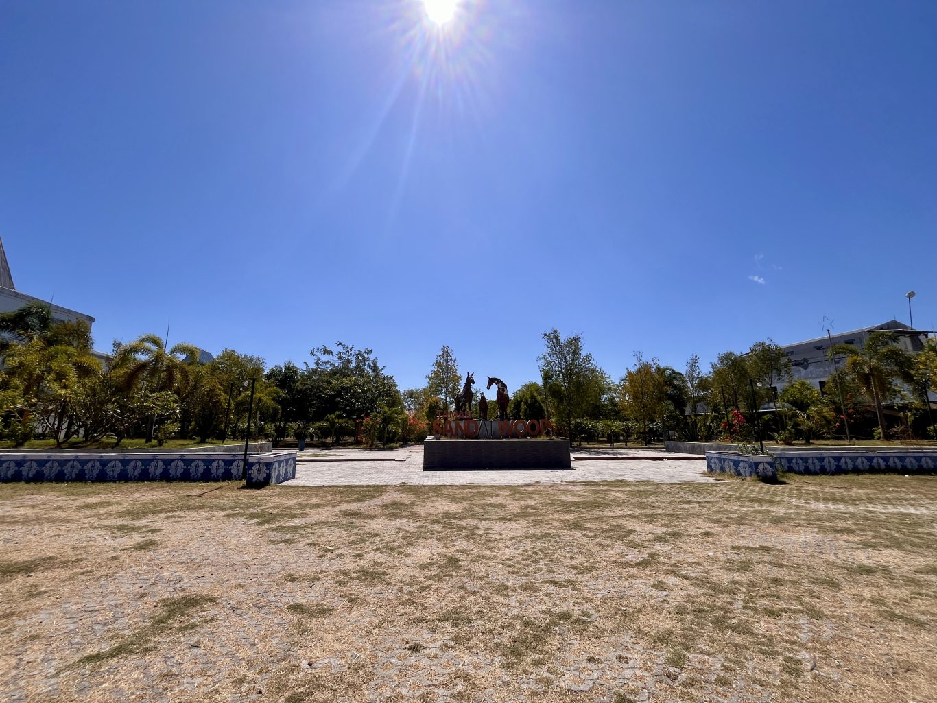 A monument featuring three dark horse statues stands prominently in Sandalwood Park, surrounded by green trees and dry grass under a bright blue sky.