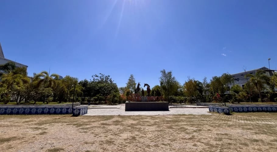 A monument featuring three dark horse statues stands prominently in Sandalwood Park, surrounded by green trees and dry grass under a bright blue sky.