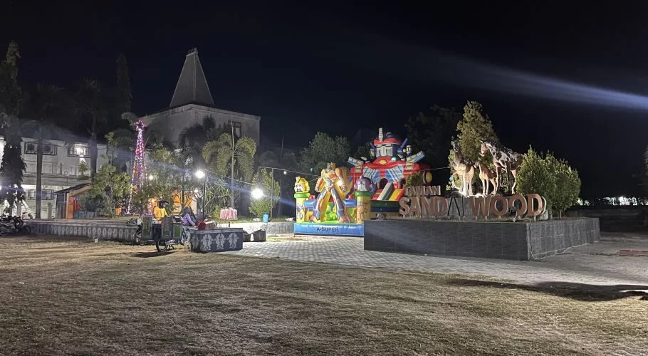 Sandalwood Park at night, illuminated by lights, shows colorful inflatable play structures, horse sculptures, and a festive tree, with visitors relaxing.