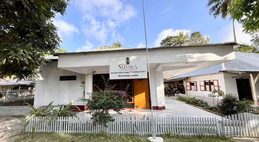 A white building with a sign for "The Sumba Foundation Malaria Microscopy Training Center" stands behind a white picket fence under a bright blue sky.