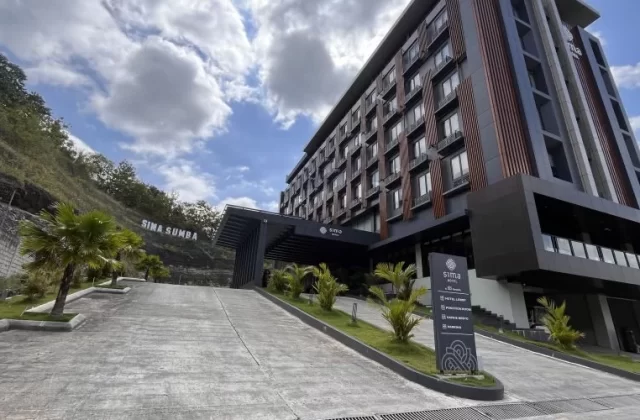 Sima Sumba Hotel, a modern multi-story building with dark grey and brown accents, stands on a green hillside with a paved entrance and palm trees.